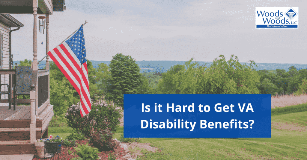 Image of an outdoor scene with a front porch of a house on the left side and an American flag attached to the front porch. Is it Hard to Get VA Disability Benefits is the title located in the bottom right corner. The Woods and Woods logo is in the top right corner. 