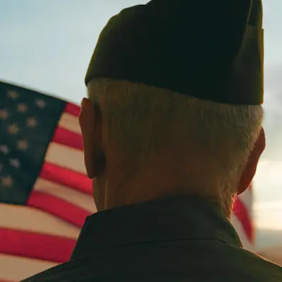 An older soldier standing with his back to the camera watching an American flag wave.